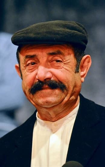 Ritratto di Benito Urgu con la coppola scura e i baffi, mentre sorride davanti a un microfono