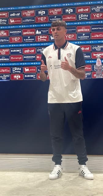 Daniel Maldini in piedi nella sala stampa del Cagliari, con la maglia bianca del club, mentre alza i pollici verso i fotografi