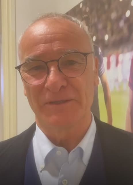 Ritratto di Claudio Ranieri, in giacca scura e camicia azzurra, davanti a una gigantografia di una partita di calcio