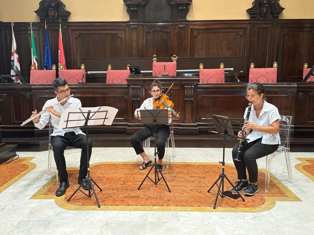 Stefano al flauto, Teresa alla viola e Angelica al clarinetto suonano seduti davanti ai leggii nell’aula magna del rettorato